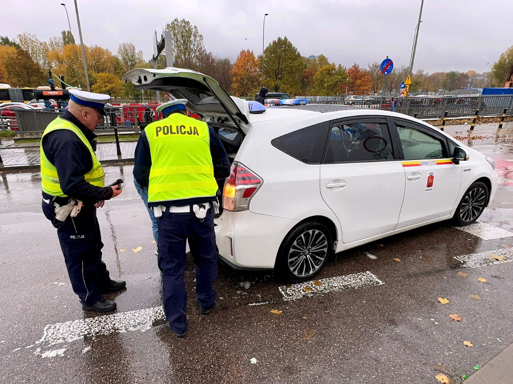 Dwaj policjanci w kamizelkach odblaskowych kontrolują białe auto na deszczowej ulicy.