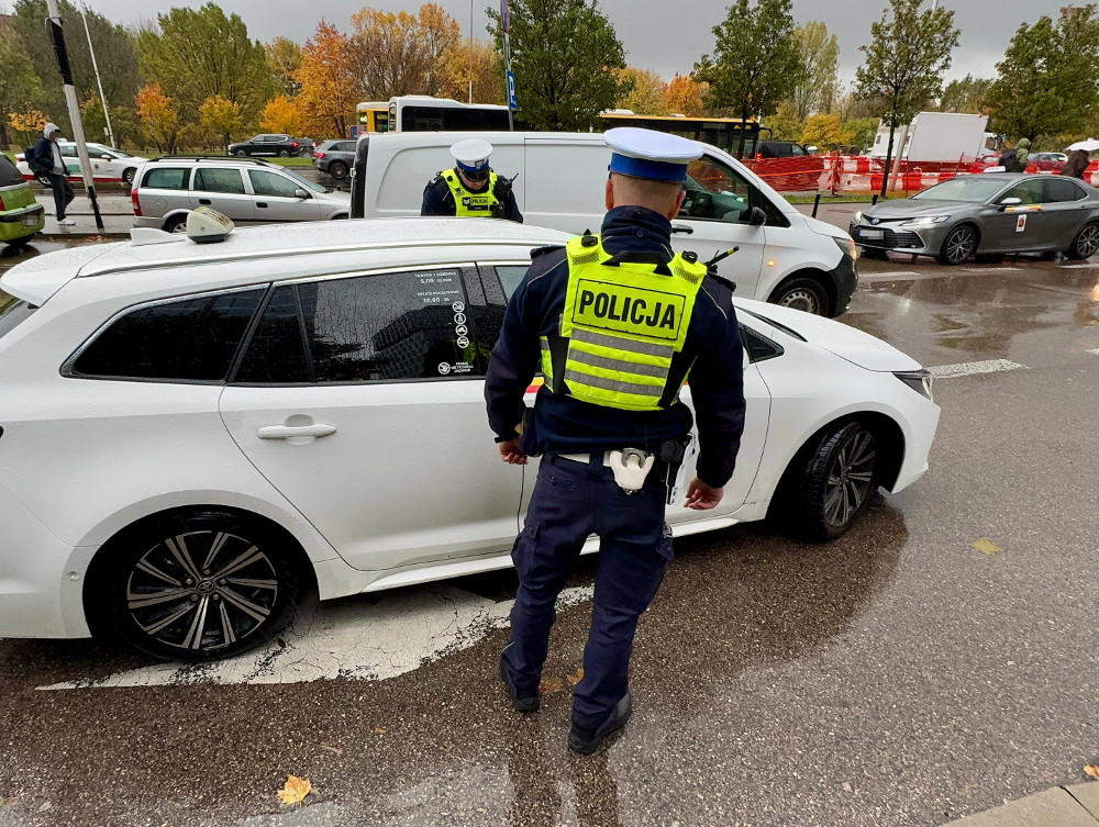 Policjant w kamizelce odblaskowej kontroluje białe auto na ruchliwej ulicy w deszczowy dzień.