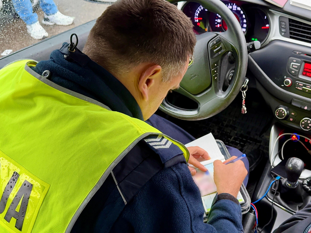Funkcjonariusz policji w odblaskowej kamizelce notuje informacje w samochodzie służbowym.