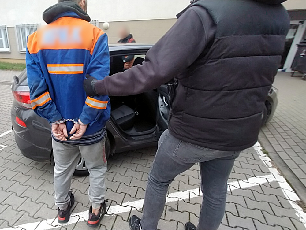 Mężczyzna w niebieskiej kurtce z pomarańczowymi elementami jest zatrzymywany przez funkcjonariusza policji przy samochodzie.