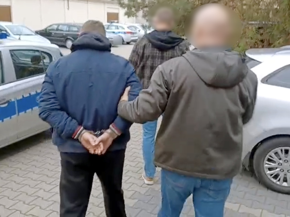 Mężczyzna w niebieskiej kurtce prowadzony przez funkcjonariusza policji na parkingu przed budynkiem.