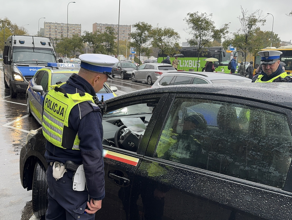Policjant w mundurze kontrolujący kierowcę w czarnym samochodzie na deszczowej ulicy, obok pojazdów policyjnych.