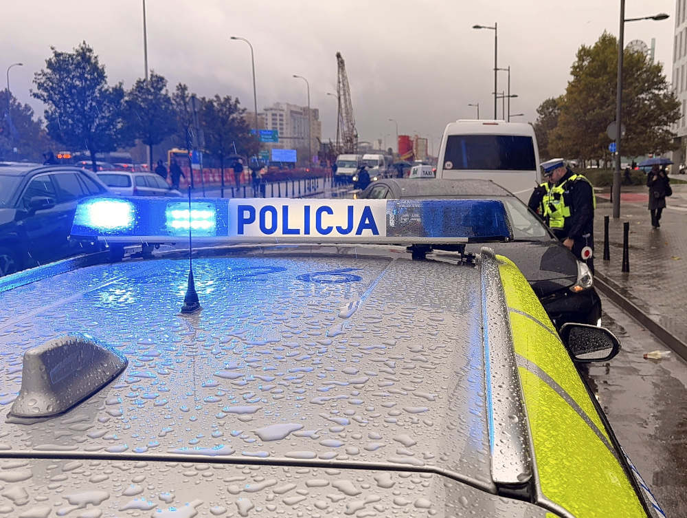 Policjant rozmawia z kierowcą przy zatrzymanym pojeździe na deszczowej ulicy, wokół stoją inne samochody.