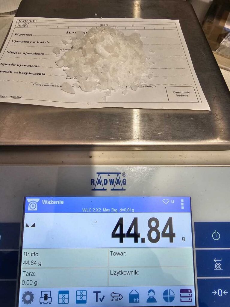 Kryształki substancji ważące 44,84 g na wadze elektronicznej, obok kartka z danymi.