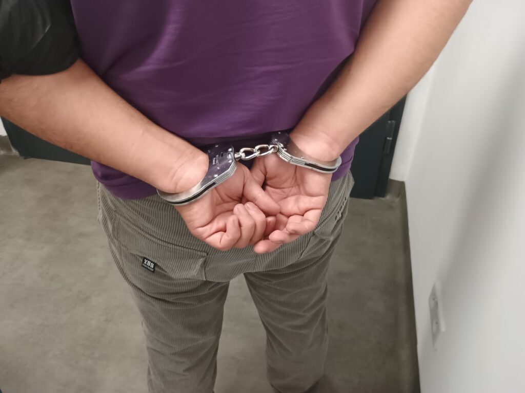 Mężczyzna w szarych spodniach ma założone kajdanki, stojąc w pomieszczeniu z betonową posadzką.