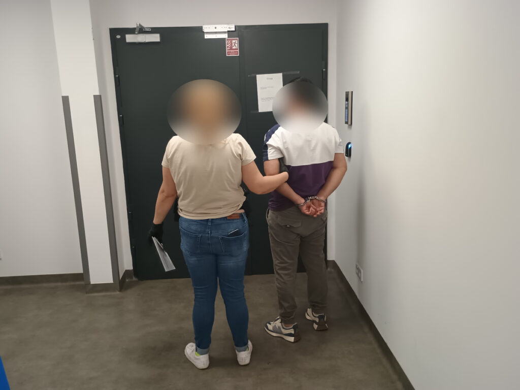 Kobieta w rękawiczkach prowadzi mężczyznę z rękami związanymi na plecach w korytarzu z szarymi ścianami.