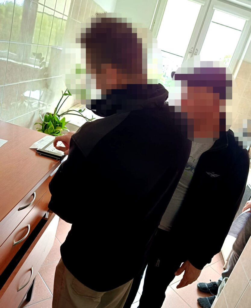 Mężczyzna w czarnej kurtce korzysta z telefonu przy drewnianym biurku, obok stoi drugi mężczyzna. W tle okno z rośliną.