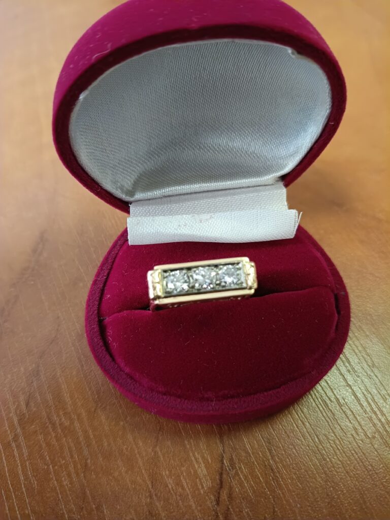 Złoty pierścionek z trzema błyszczącymi diamentami, umieszczony w czerwonym, welurowym pudełku na tle drewnianego blatu.