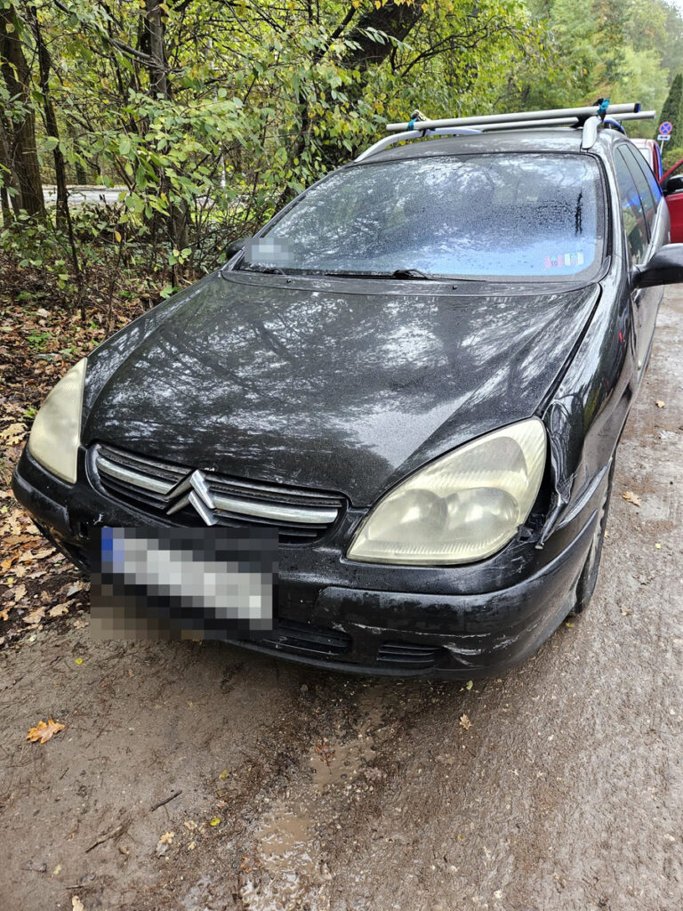 Czarny samochód osobowy zaparkowany na błotnistej drodze, otoczony przez drzewa i opadłe liście.