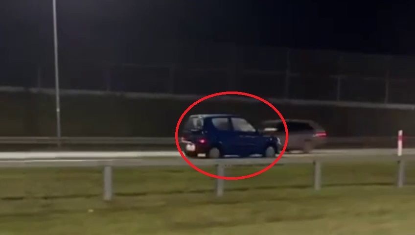 Niebieski samochód osobowy porusza się wzdłuż drogi nocą obok ciemnego pojazdu.