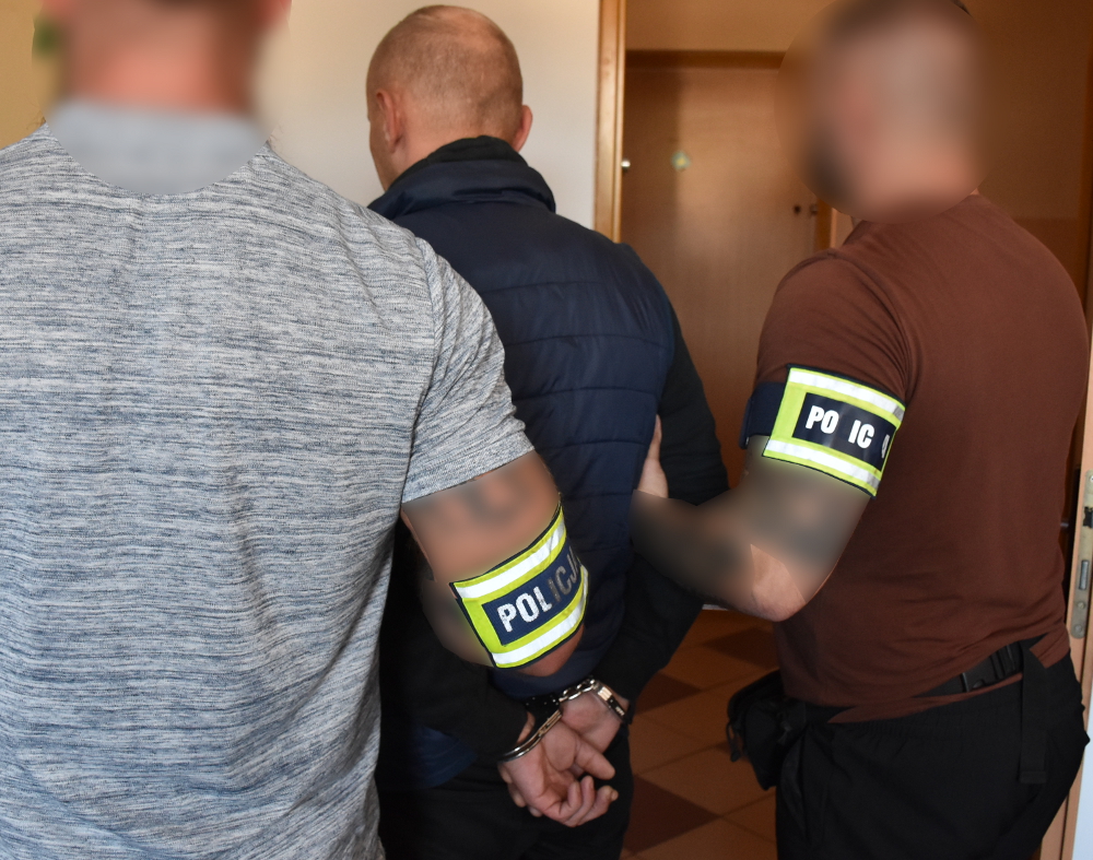 Trzej policjanci w mundurach z odznakami prowadzą mężczyznę w kajdankach w korytarzu budynku.