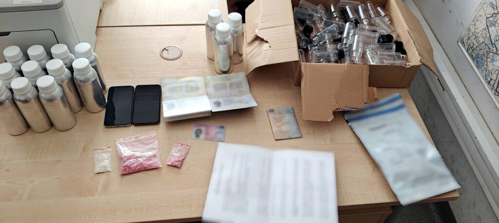 Na blacie biurka znajdują się butelki, dwa telefony, pakunki z substancjami oraz dokumenty tożsamości.