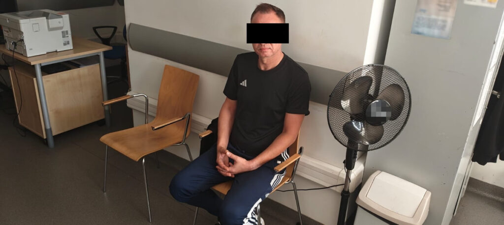 Mężczyzna w czarnej koszulce siedzący na krześle w pomieszczeniu z biurkiem i wentylatorem.