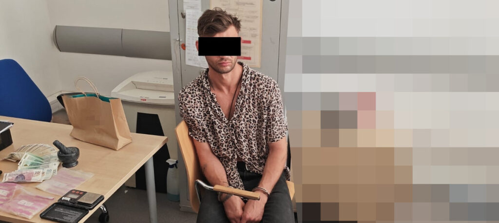 Mężczyzna w koszuli w panterkę siedzi na krześle w pomieszczeniu biurowym, z rękami związanymi metalowym łańcuchem. Na stole leżą pieniądze i przedmioty osobiste.