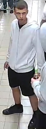 Młody mężczyzna w białej bluzie z kapturem stoi w sklepie, patrząc w stronę kamery. Ma na sobie czarne szorty i buty sportowe.