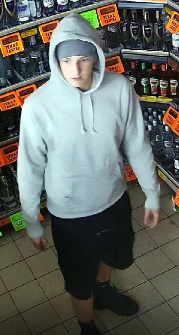 Młody mężczyzna w szarym dresie z kapturem stoi w sklepie, otoczony półkami z napojami.