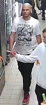 Mężczyzna w białej koszulce z nadrukiem przechodzi obok sklepu w ruchliwym korytarzu handlowym.