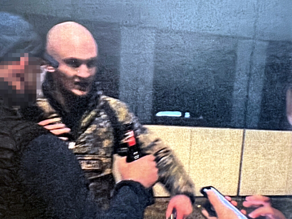 Mężczyzna w mundurze wojskowym rozmawia z innymi osobami w pomieszczeniu z kafelkami.