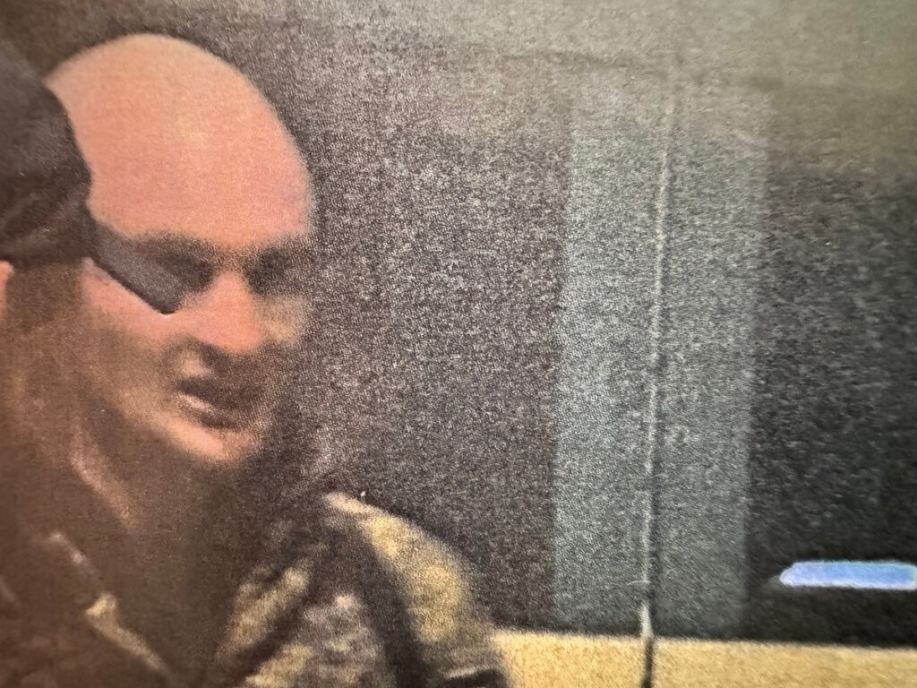 Mężczyzna z częściowo łysą głową stoi w towarzystwie innych osób, w tle widoczne zamglone kontury budynków.