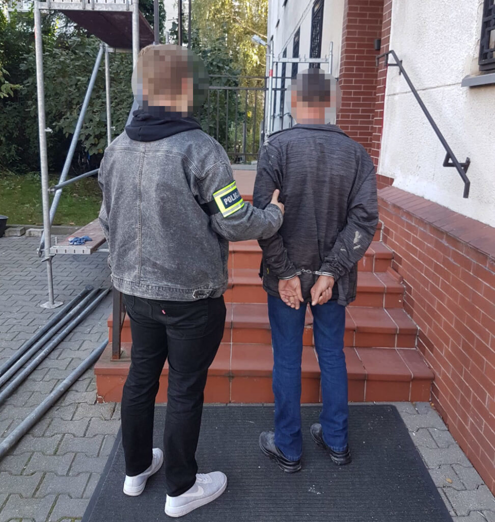Funkcjonariusz policji w dżinsowej kurtce prowadzi zatrzymanego mężczyznę na schodach budynku.