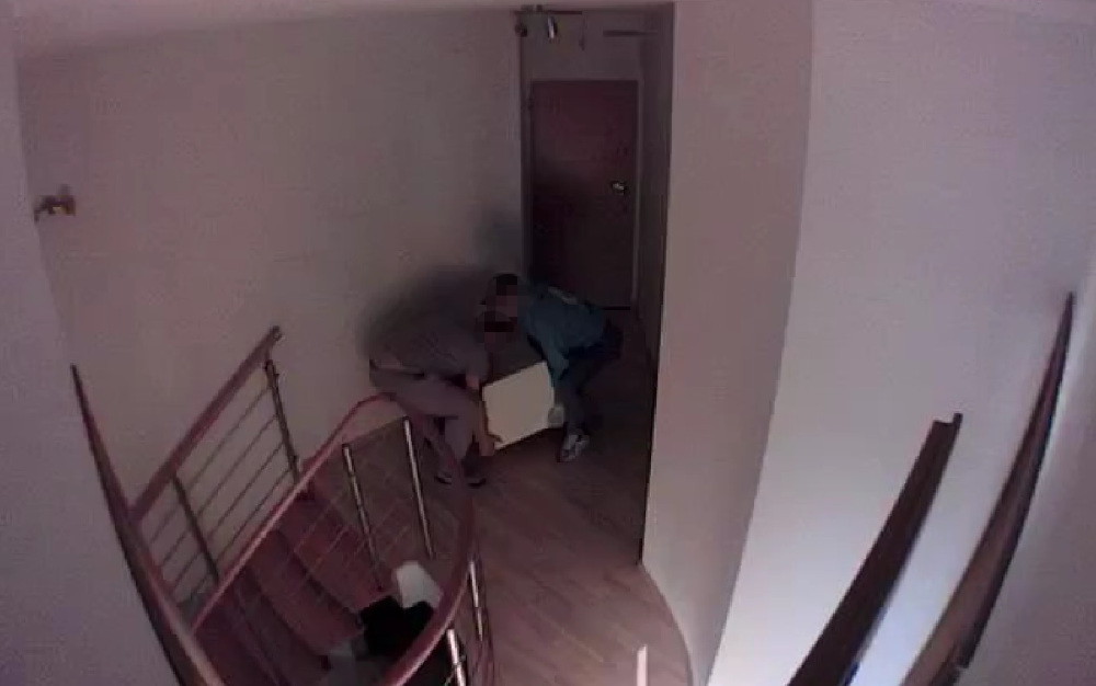 Dwaj mężczyźni przenoszą szafkę w wąskim korytarzu, obok schodów i zamkniętych drzwi.