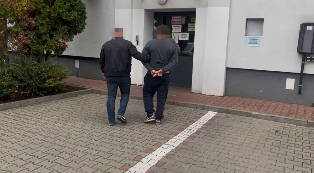 Mężczyzna w dżinsach prowadzony przez policjanta w kierunku budynku z przeszklonym wejściem.