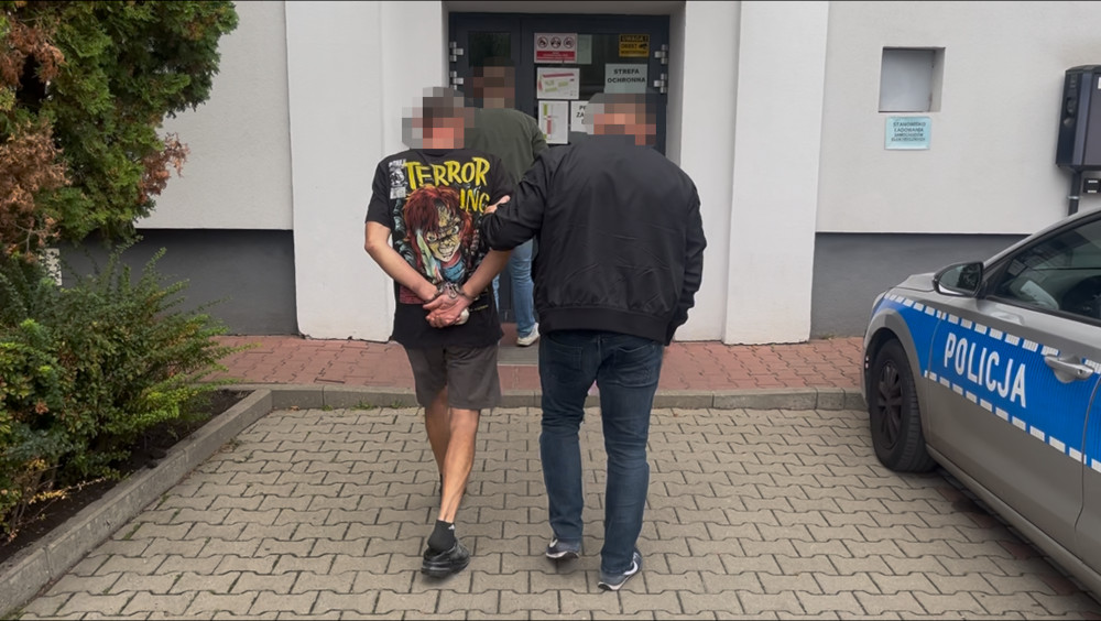 Mężczyzna w koszulce z nadrukiem prowadzony przez policjanta przed budynkiem z oznaczeniem "strefa ochronna".