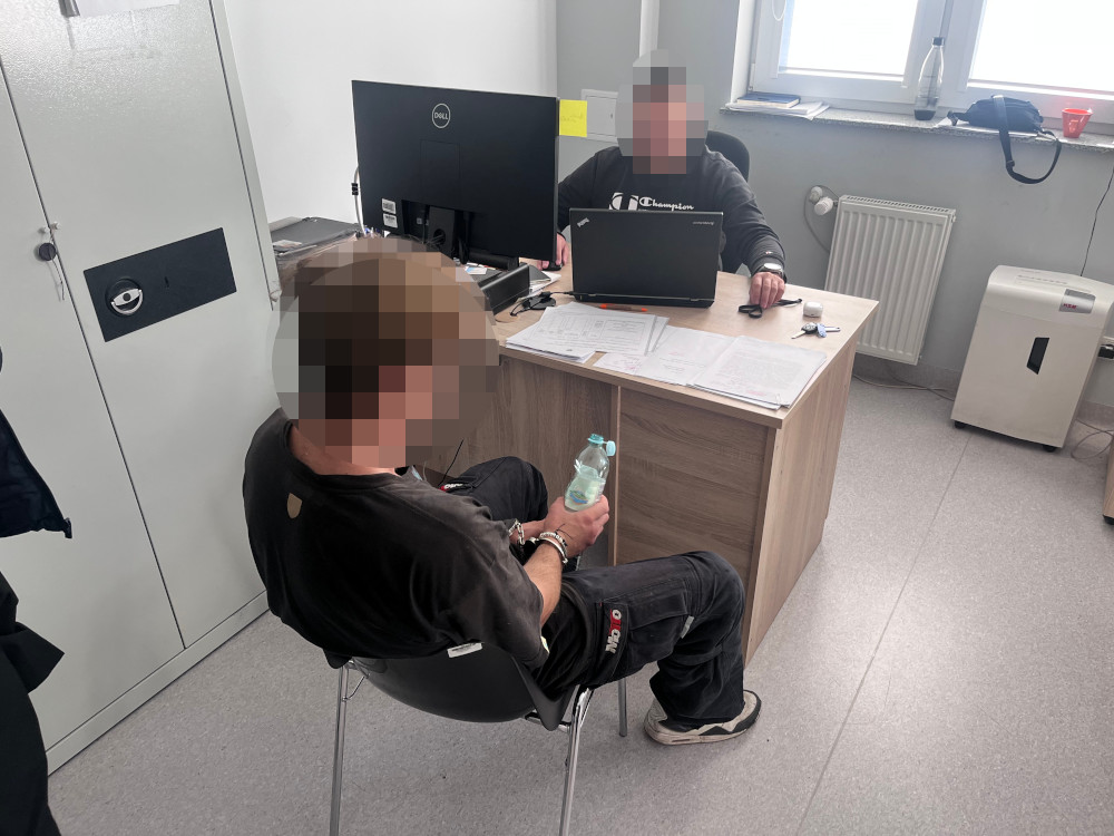 Mężczyzna siedzący na krześle trzyma butelkę wody, podczas gdy inny mężczyzna pracuje na laptopie przy biurku.