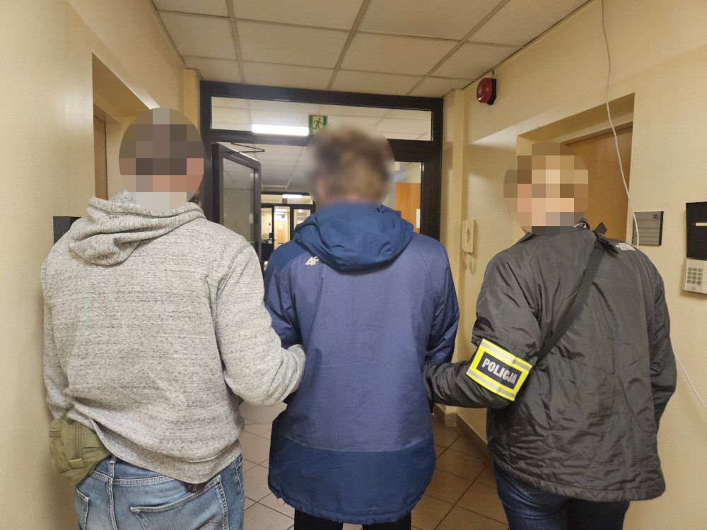 Trzech mężczyzn idących korytarzem, jeden z nich w niebieskiej kurtce, eskortowany przez funkcjonariusza z opaską "POLICJA".