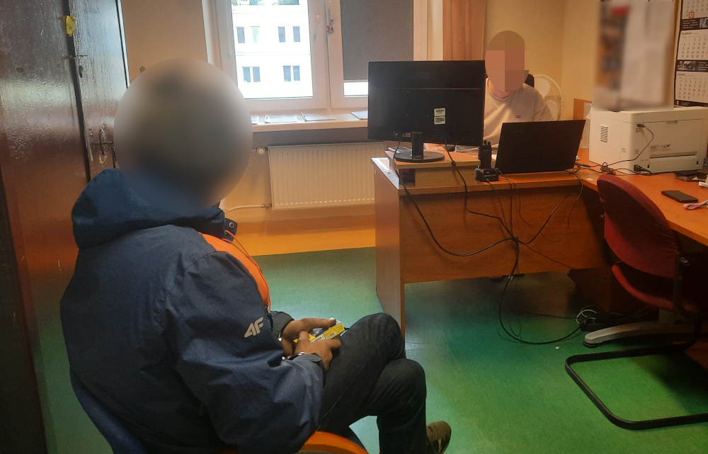 Mężczyzna w niebieskiej kurtce siedzi na krześle, trzymając w ręku telefon, podczas rozmowy z innym mężczyzną przy biurku.