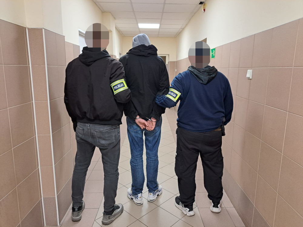 Trzech funkcjonariuszy policji prowadzi zatrzymanego mężczyznę korytarzem w budynku.
