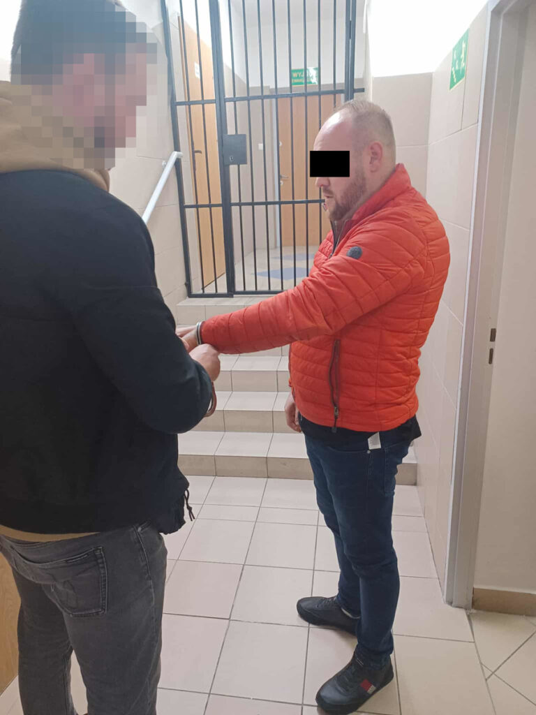 Mężczyzna w czerwonej kurtce stoi na schodach, podczas gdy inny mężczyzna zakłada mu kajdanki w budynku z kratami.