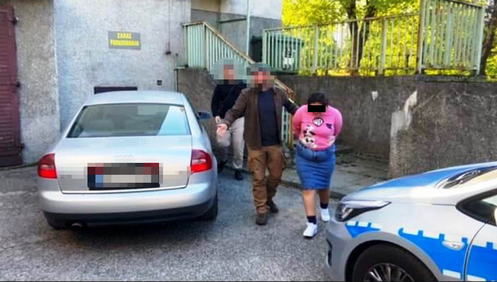 Mężczyzna w brązowej kurtce prowadzi kobietę w różowej koszulce na terenie parkingu, obok zaparkowanych samochodów.