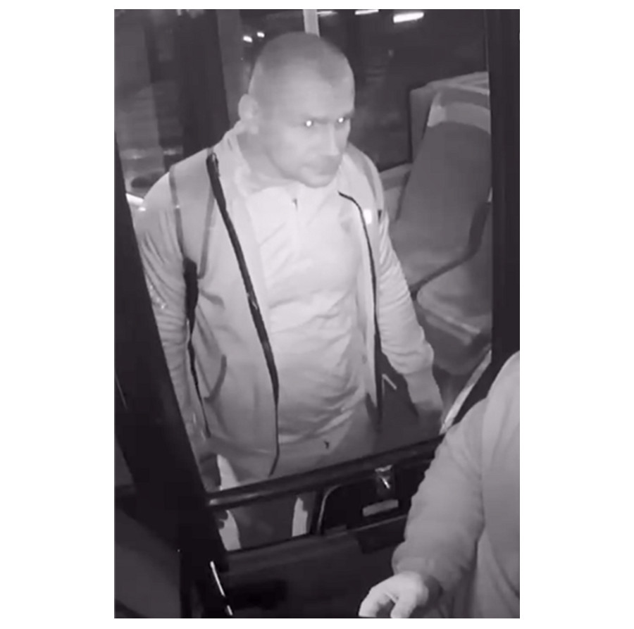 Mężczyzna w jasnym dresie wchodzi do autobusu, stojąc blisko drzwi, obok drugiego mężczyzny w ciemnym ubraniu.
