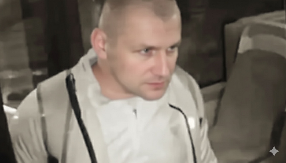 Mężczyzna w białej bluzie z zamkiem, z krótkimi włosami, stoi w jasnym pomieszczeniu z meblami w tle.
