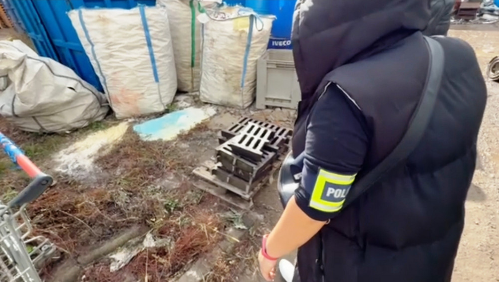 Funkcjonariusz policji bada teren w pobliżu worków z odpadami i paletą na zarośniętej trawie.