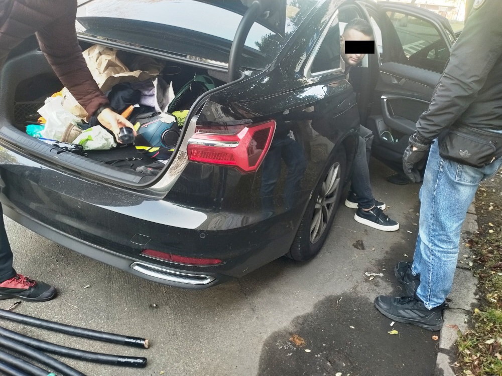 Mężczyzna przeszukujący bagażnik ciemnego samochodu osobowego, obok stoi inny mężczyzna.