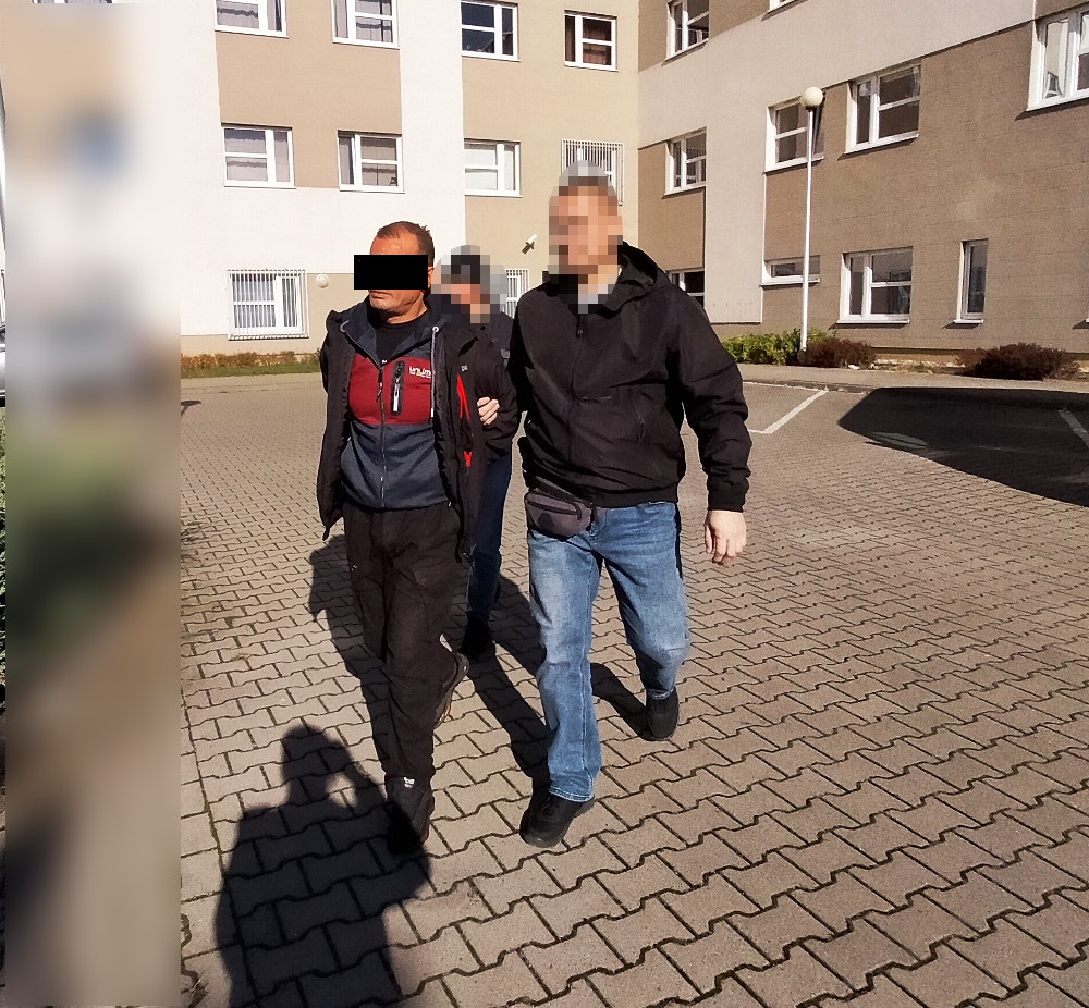 Mężczyzna w ciemnym ubraniu prowadzony przez funkcjonariusza w jasnej kurtce na parkingu przed budynkiem mieszkalnym.