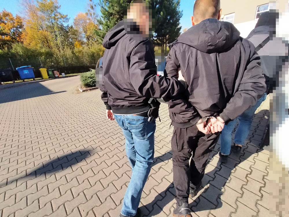 Mężczyzna w dżinsach prowadzony przez funkcjonariusza policji na terenie osiedla, w tle kosze na śmieci.