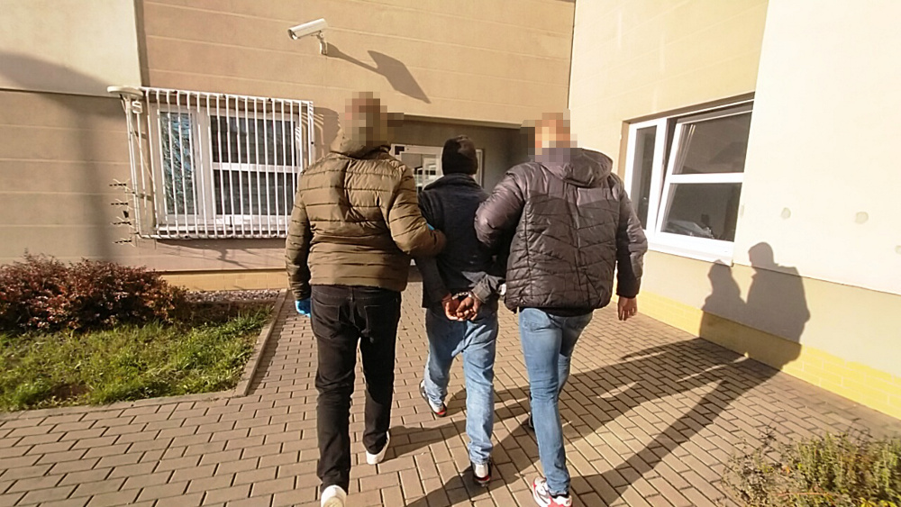 Trzech mężczyzn, z których jeden jest w kajdankach, idzie wzdłuż budynku, prowadzeni przez dwóch innych.