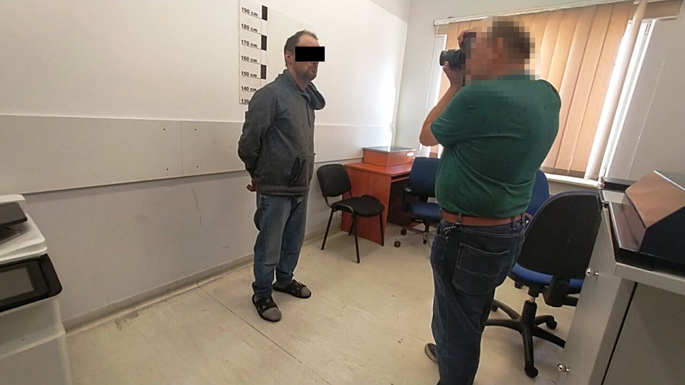 Mężczyzna w szarej kurtce stoi na tle miary wzrostu, podczas gdy fotograf robi mu zdjęcie w pomieszczeniu z biurkami.