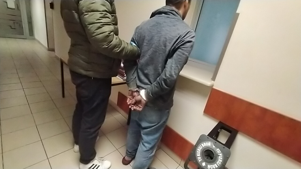 Mężczyzna w szarej bluzie stoi z rękami z tyłu, trzymany przez funkcjonariusza w korytarzu budynku.