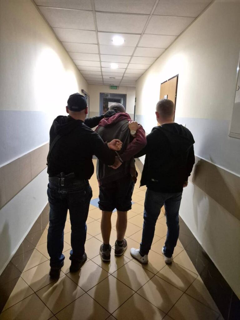 Trzech mężczyzn prowadzi osobę z rękami skrępowanymi z tyłu korytarzem w budynku.