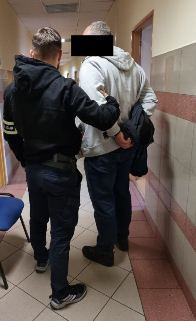 Policjant w mundurze prowadzi mężczyznę w szarym dresie przez korytarz w budynku administracyjnym.