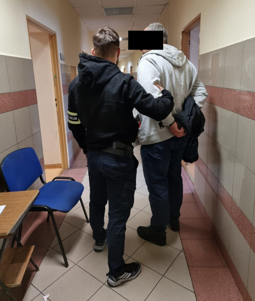 Funkcjonariusz policji prowadzi mężczyznę w szarym dresie korytarzem w budynku, trzymając go za ramię.
