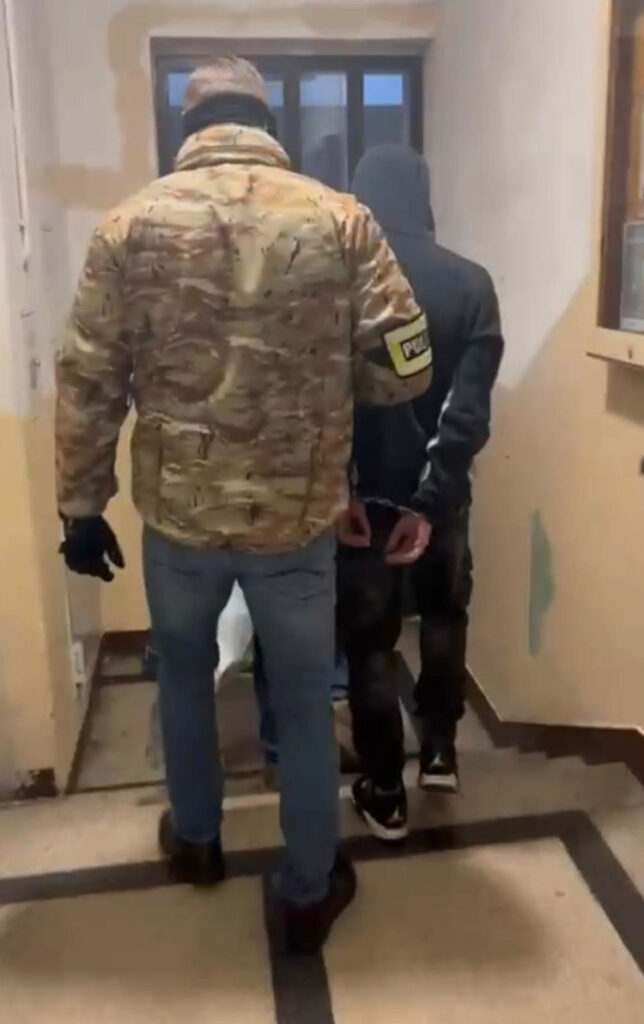 Funkcjonariusz w kamizelce policyjnej prowadzi zatrzymanego mężczyznę w ciemnym dresie przez korytarz.