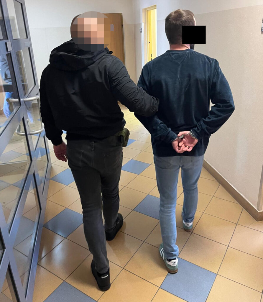 Funkcjonariusz policji prowadzi mężczyznę z rękami z tyłu w korytarzu budynku.