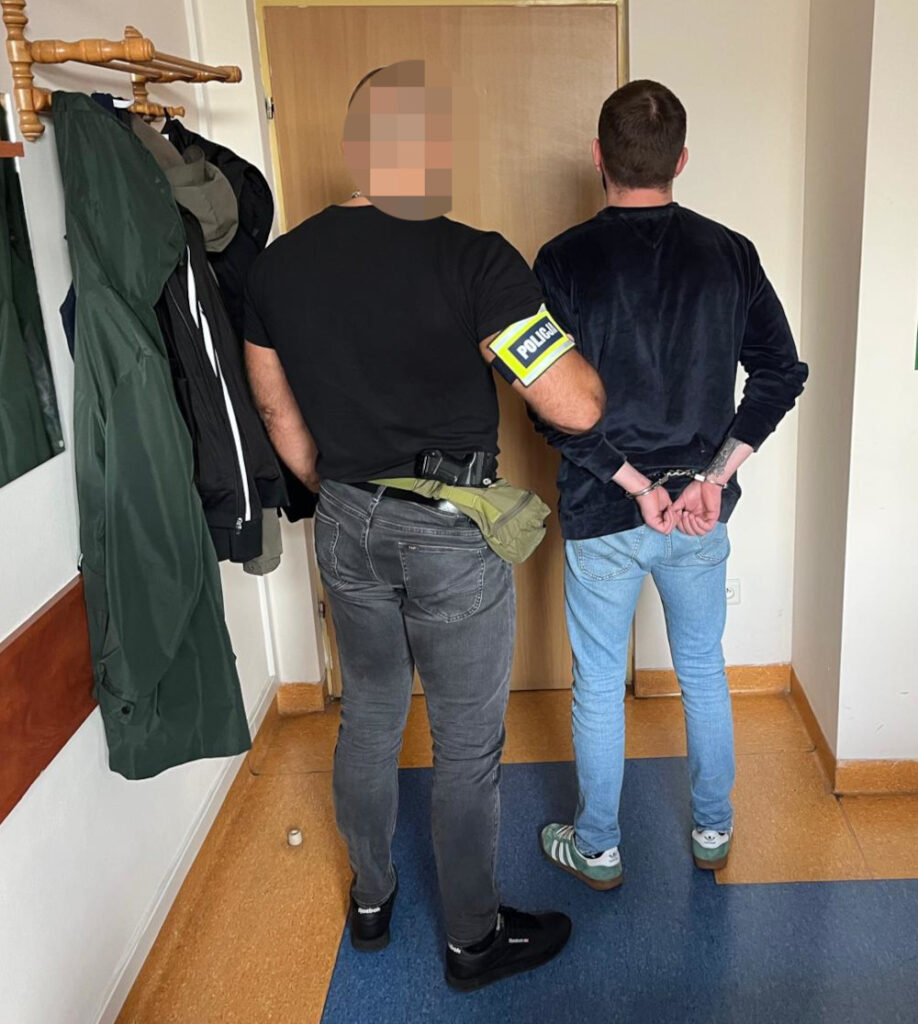 Funkcjonariusz policji w czarnym t-shircie eskortuje mężczyznę w kajdankach w korytarzu budynku.