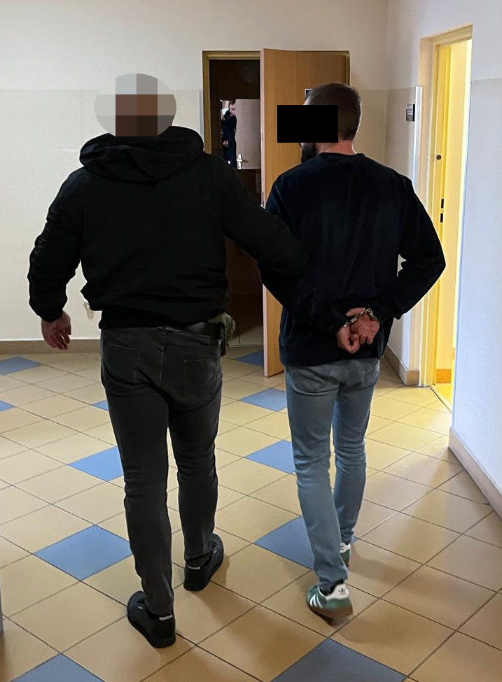 Funkcjonariusz policji prowadzi zatrzymanego mężczyznę korytarzem w budynku.