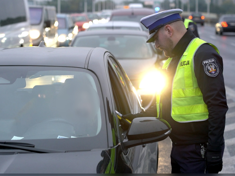 Policjant w kamizelce odblaskowej kontroluje kierowcę w samochodzie osobowym na zatłoczonej drodze.